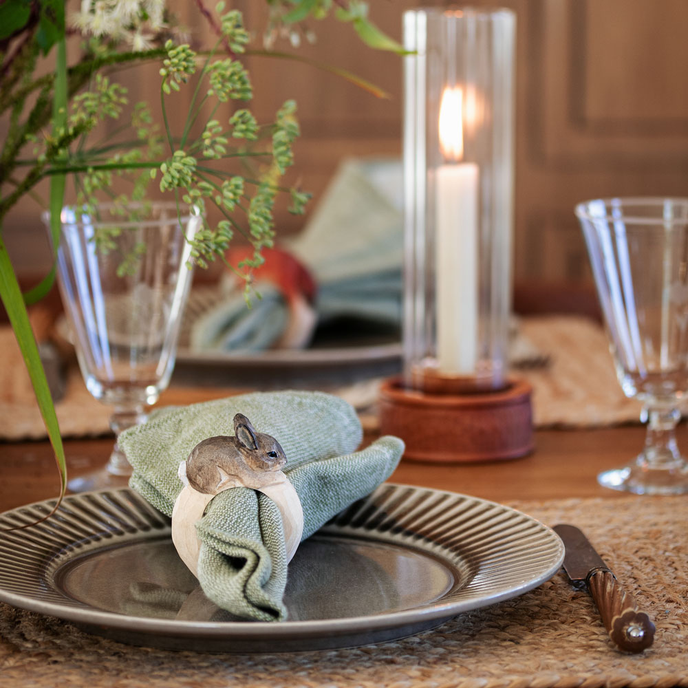 Une table dressée avec le Rond de serviette – Lapin Mini autour d’une serviette verte sur une assiette, entouré de verres et d’une bougie allumée.