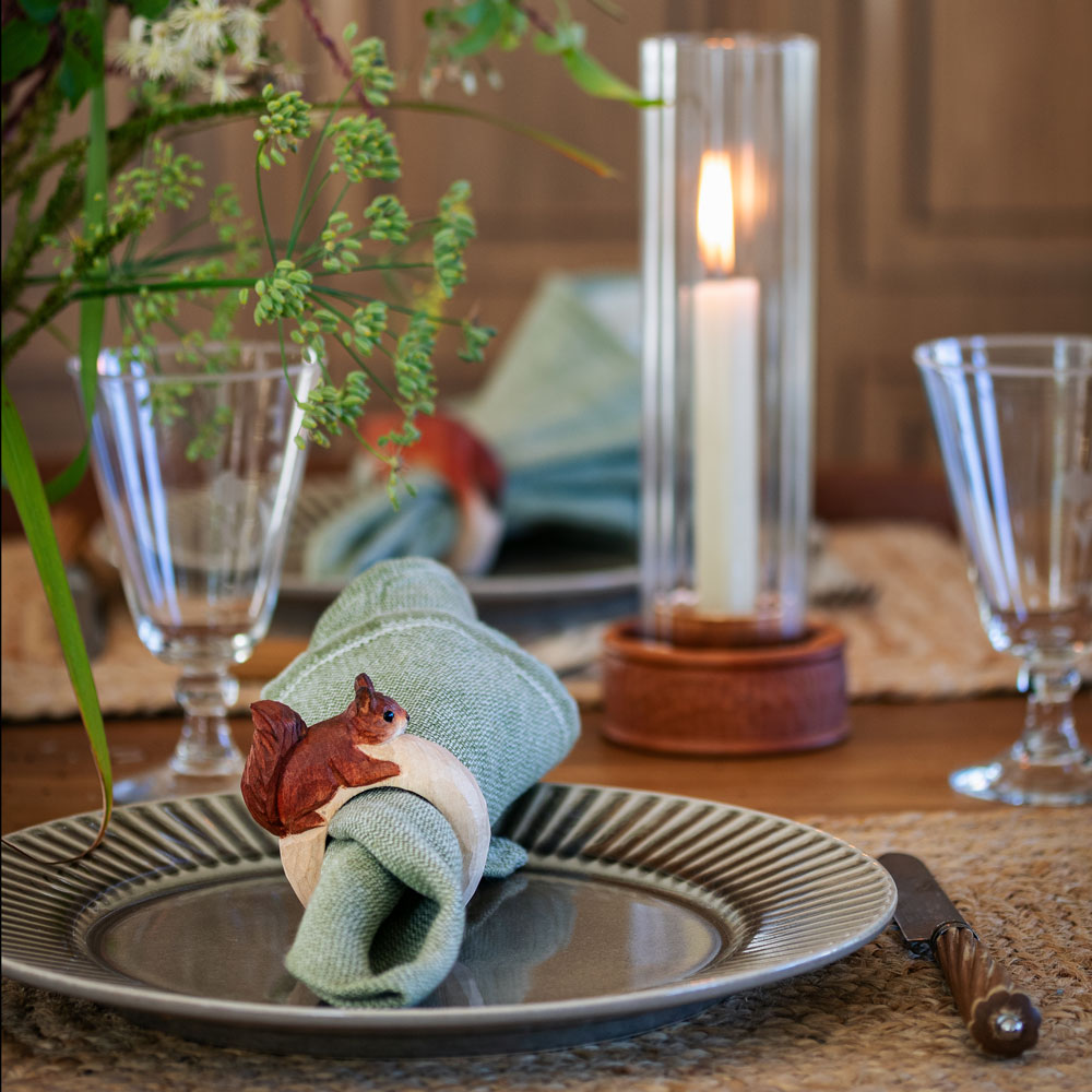 Une table dressée avec le Rond de serviette – Écureuil Mini tenant une serviette verte sur une assiette, entouré de verres, de feuillage et d’une bougie allumée.