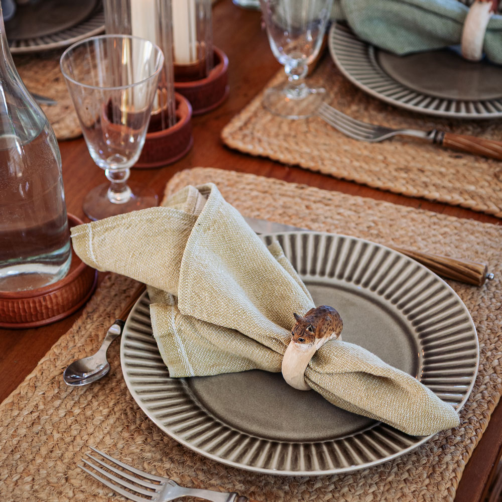 Une table dressée avec le Rond de serviette – Lynx Mini autour d’une serviette beige sur une assiette, entouré de verres et d’une carafe d’eau.