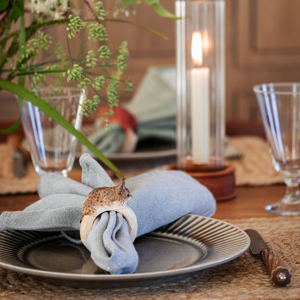 Une table dressée avec le Rond de serviette – Lynx Mini autour d’une serviette bleue sur une assiette, avec des verres et une bougie allumée en arrière-plan.