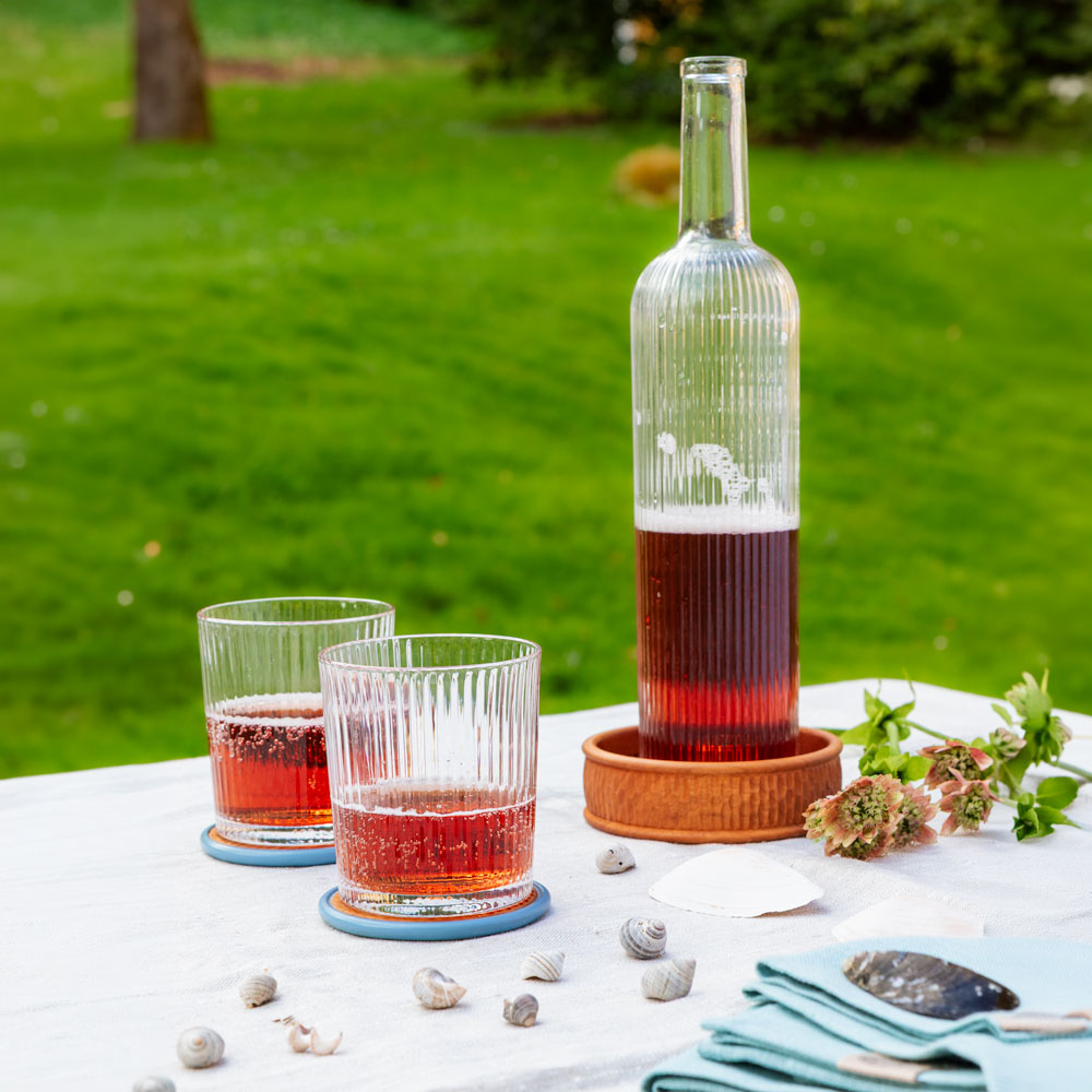 Dessous de bouteille de la série Onde avec bouteille en verre, verres, coquillages et textiles bleu clair sur une table de jardin.