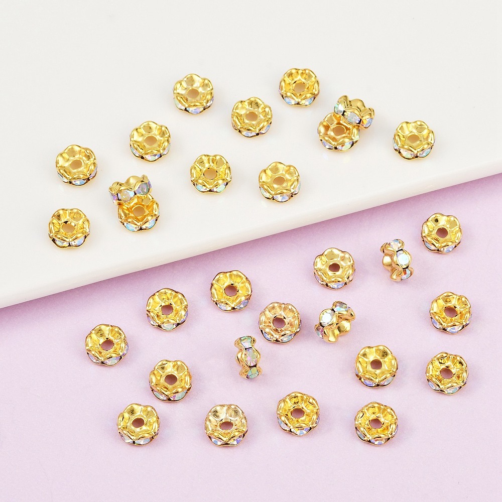 Strassrondell Clear AB, rund 6 mm rhinestone-rondell i gyllene metallfärg med skimrande AB-stenar, utspridda på vit och rosa bakgrund i närbild.