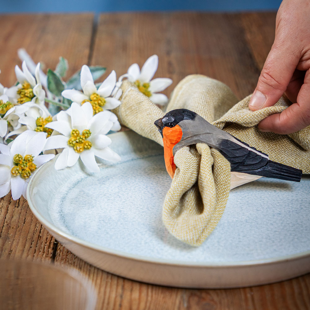 Rond de Serviette Bouvreuil Pivoine autour d’une serviette en tissu sur une table dressée avec des fleurs blanches.