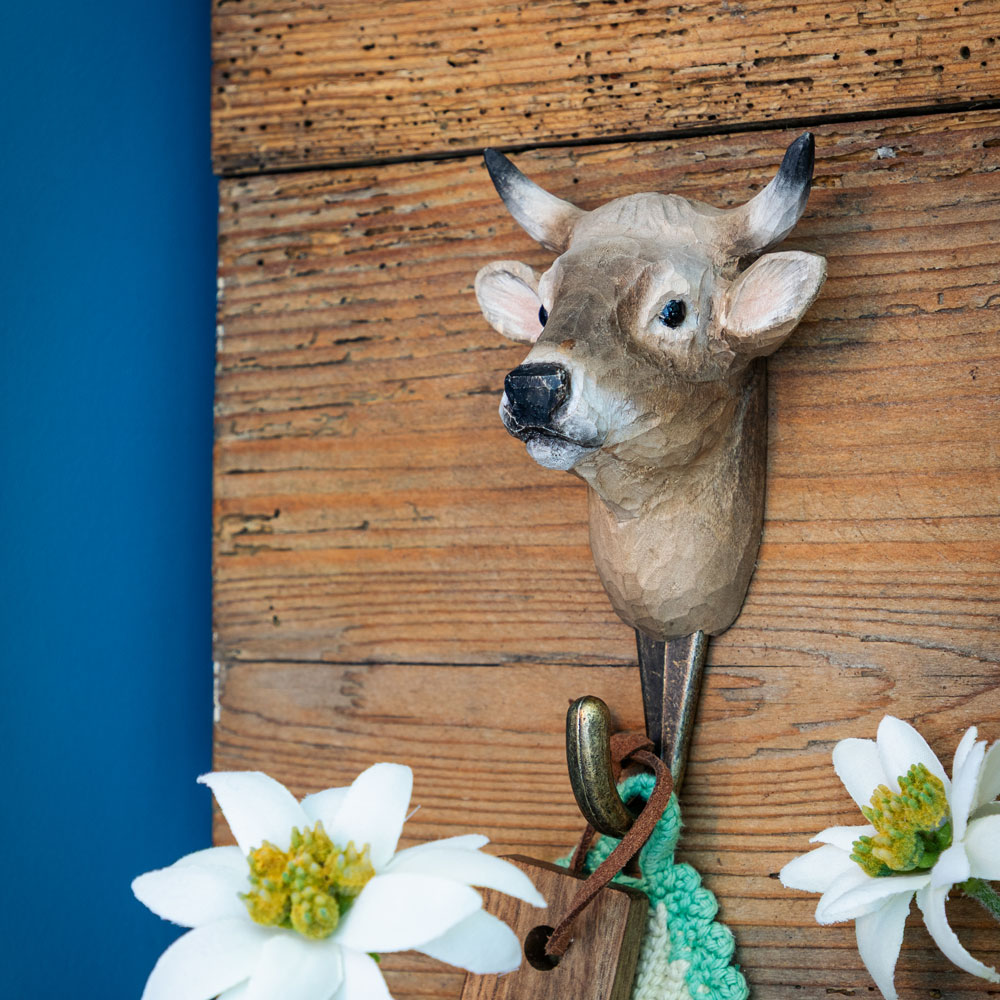 Patère sculptée à la main avec vache alpine fixée sur un mur en bois rustique, à côté d’une manique tricotée et d’une peau de mouton.