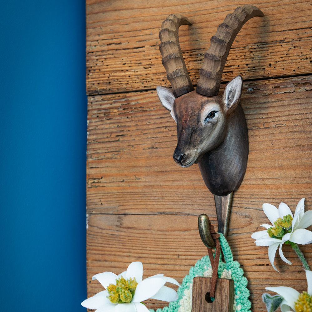 Patère avec bouquetin des Alpes sculpté à la main fixée sur un mur en bois, à côté d’une peau de mouton et d’une planche ronde en bois.