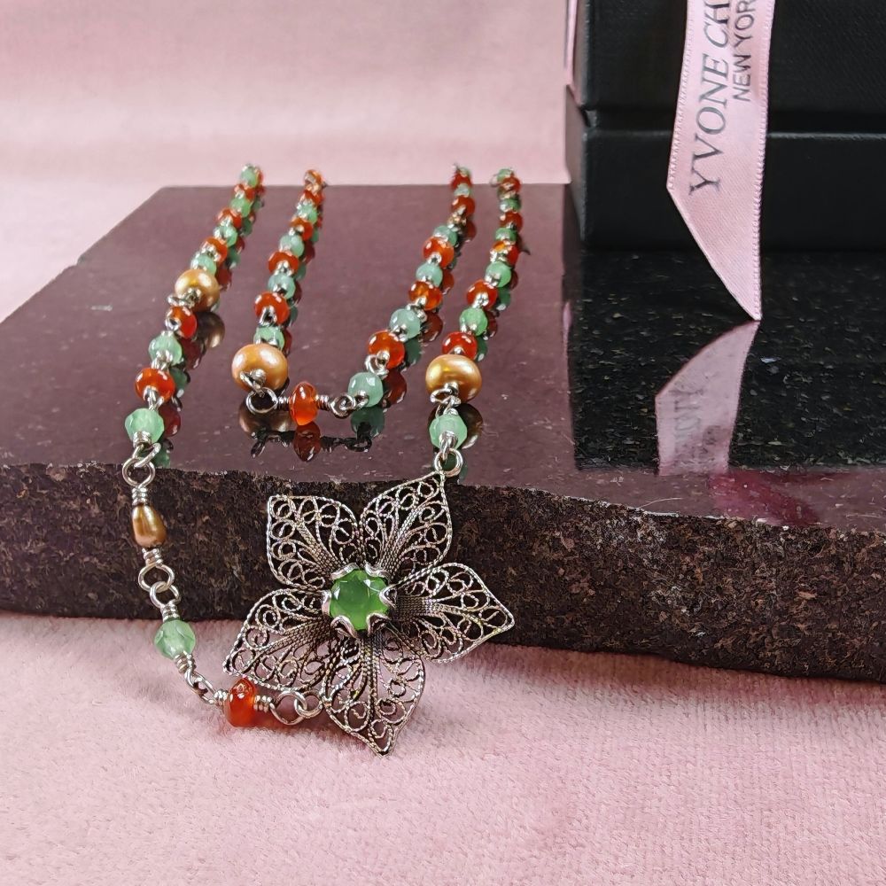 Yvone Christa Menage Long Twisted halsband, långt halsband i återvunnet sterling silver med karneol, grön jade och ljusbruna sötvattenspärlor. Halsbandet ligger draperat på en rosa duk och svart stenskiva, med ett blomformat filigranhänge med grön sten i närbild.