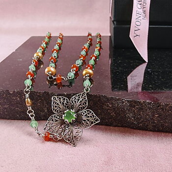 Yvone Christa Menage Long Twisted halsband, långt halsband i återvunnet sterling silver med karneol, grön jade och ljusbruna sötvattenspärlor. Halsbandet ligger draperat på en rosa duk och svart stenskiva, med ett blomformat filigranhänge med grön sten i närbild.