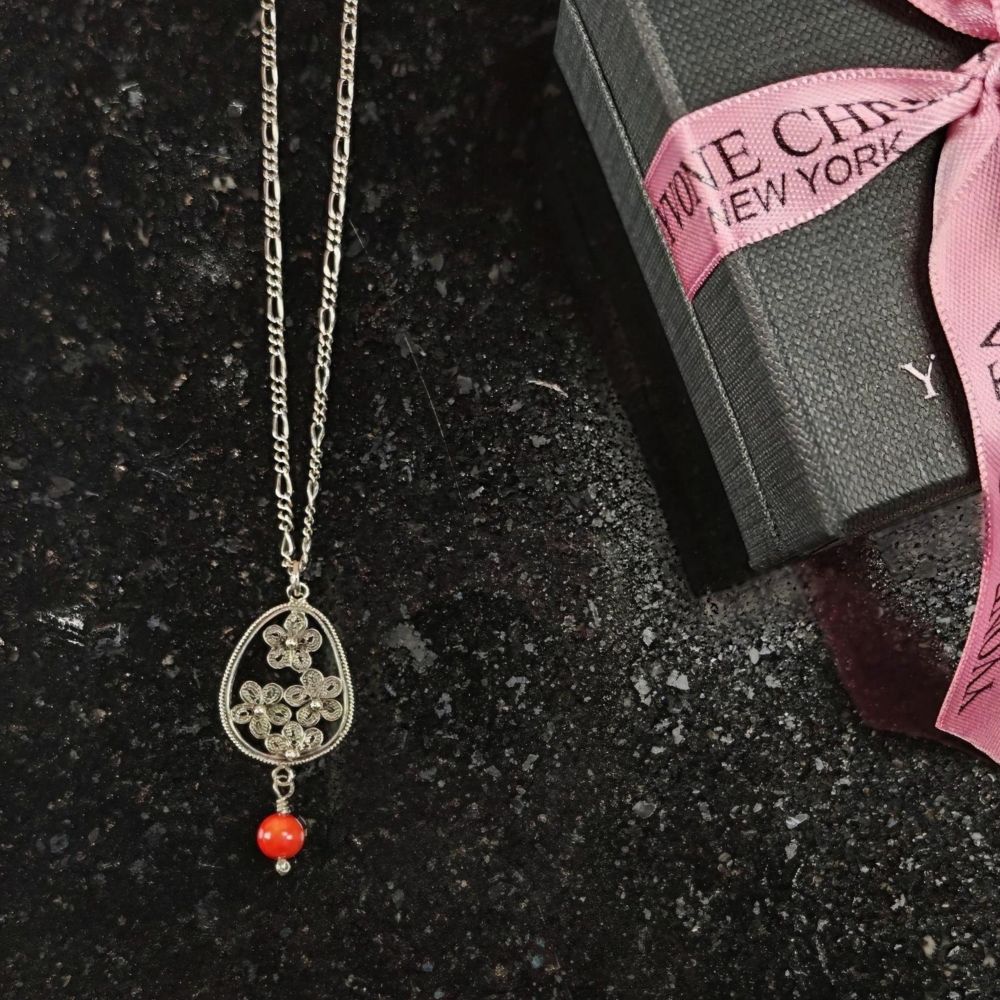 Yvone Christa Menage halsband i sterling silver med droppformat filigranhänge och röd korall. Halsbandet ligger på en mörk stenskiva bredvid en svart presentask med rosa band.