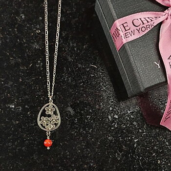 Yvone Christa Menage halsband i sterling silver med droppformat filigranhänge och röd korall. Halsbandet ligger på en mörk stenskiva bredvid en svart presentask med rosa band.
