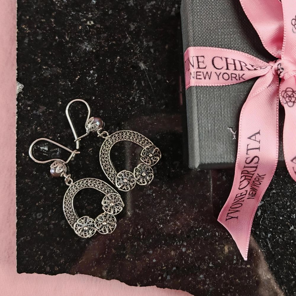 Yvone Christa Tiny Hanging Bouquet of Flower örhängen i filigransilver med svart pärla. Örhängena ligger på en mörk stenskiva bredvid en svart presentask med rosa Yvone Christa-band, mot rosa bakgrund.