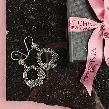 Yvone Christa Tiny Hanging Bouquet of Flower örhängen i filigransilver med svart pärla. Örhängena ligger på en mörk stenskiva bredvid en svart presentask med rosa Yvone Christa-band, mot rosa bakgrund.