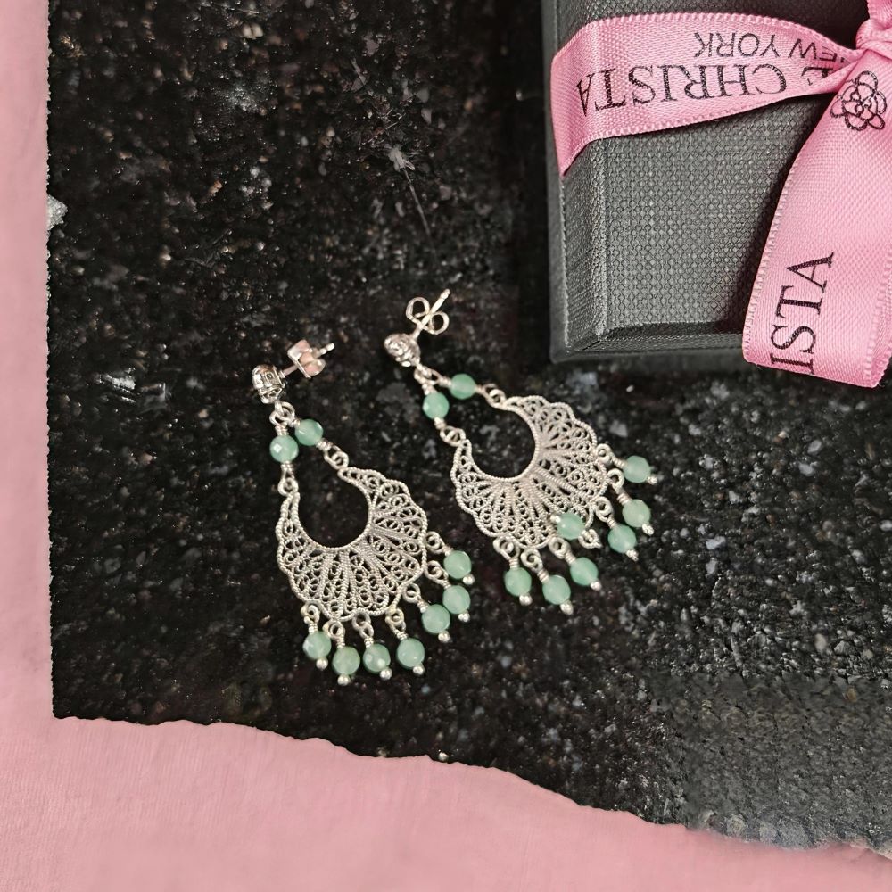 Yvone Christa Menage Tulip Post örhängen i filigransilver med gröna jadepärlor och liten CZ-sten. Örhängena ligger på en mörk stenskiva bredvid en svart presentask med rosa Yvone Christa-band, mot rosa bakgrund.
