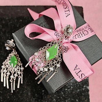 Yvone Christa Menage Flower Post örhängen i filigransilver med limegrön kristall och beige pärlor. Ett örhänge ligger på en mörk stenskiva och det andra hänger på en svart presentask med rosa Yvone Christa-band, mot rosa bakgrund.