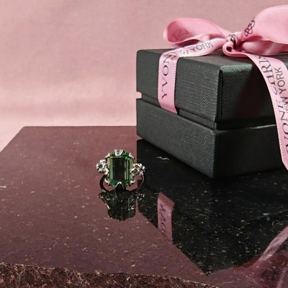 Yvone Christa Menage Square ring i sterling silver med stor fyrkantigt slipad grön Aqua Lemurian-sten, placerad på en mörk stenskiva bredvid en svart presentask med rosa band.
