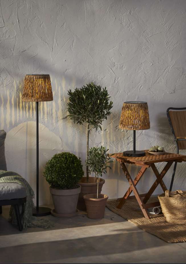 Flätverk solar rattan floor lamp glowing on a patio beside olive trees, a boxwood ball and the matching table lamp