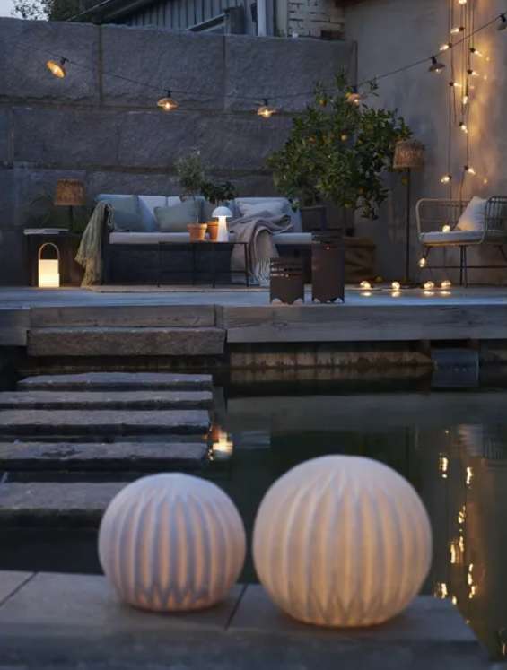 Cesta rattan solar lamp glowing on a dusk patio with string lights and outdoor sofa by a pool, seen past two round globe lamps