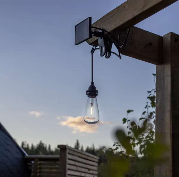 Close-up of the solar panel and clip fixed to a pergola beam, the bare filament bulb glowing against a blue evening sky
