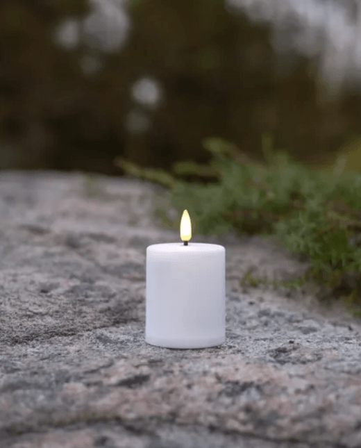 Ett vitt batteridrivet LED-blockljus med verklighetstrogen låga, stående utomhus på en grå sten i naturen.