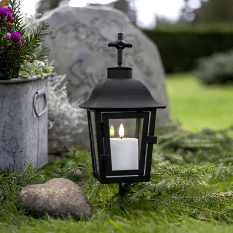 Svart gravlykta i metall med ett vitt batteridrivet LED-ljus inuti, placerad på gräs bredvid en gravsten och en hjärtformad sten.