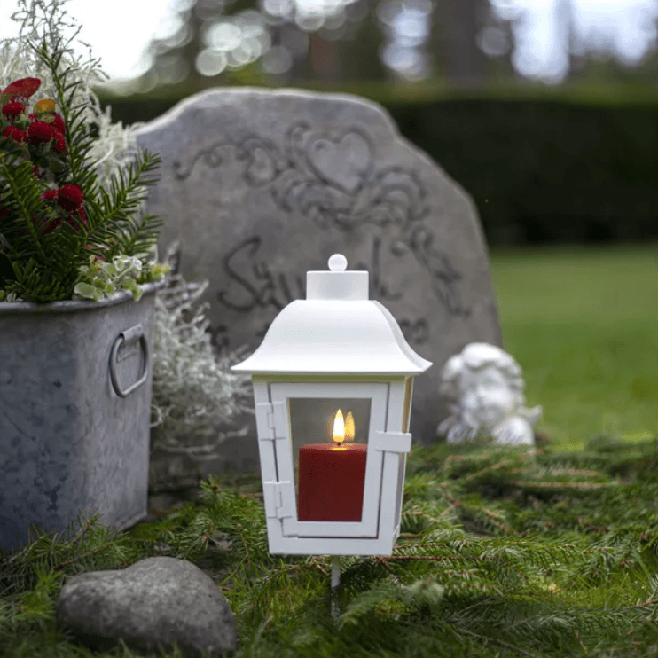 Vit gravlykta i metall med ett rött batteridrivet LED-ljus inuti, placerad vid en gravsten bredvid en hjärtformad sten och en liten ängel.