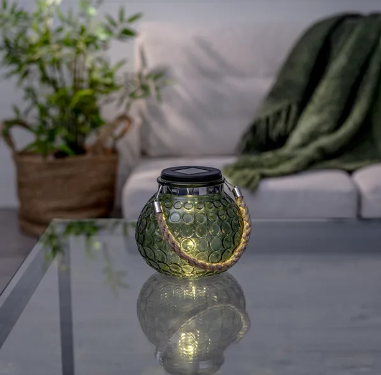 Green decorative glass solar lantern with a rope handle resting on a glass table indoors.