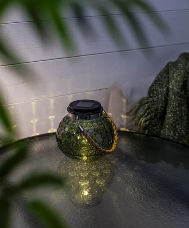 Glowing green glass solar lantern casting warm patterned light reflections on a dark outdoor table at night.