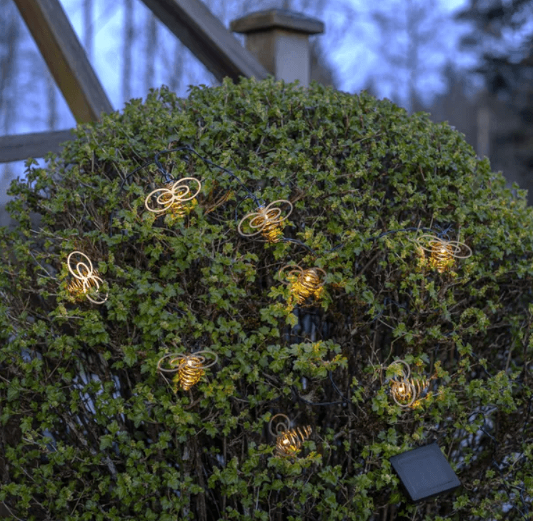 Solar-powered bee shaped string lights illuminating a green garden bush at dusk.
