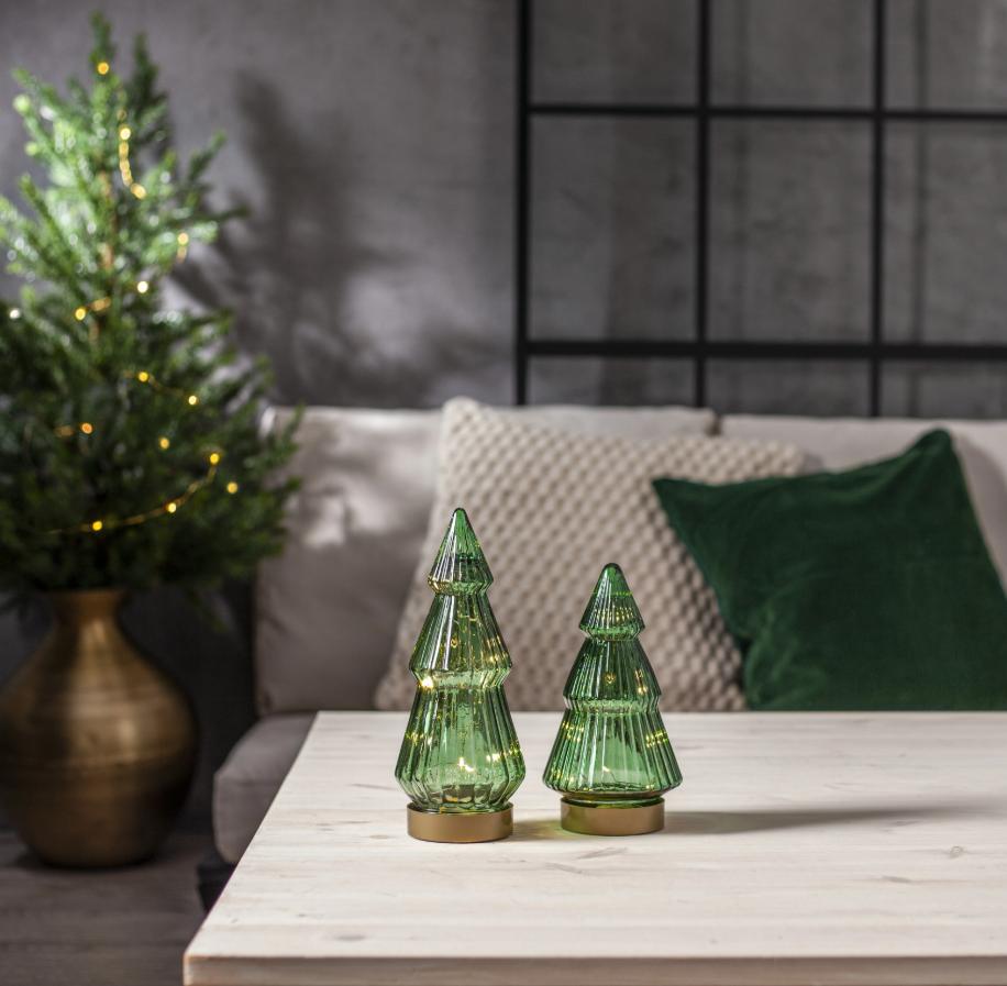 vå gröna julgranar i glas med stämningsfull LED-belysning i olika storlekar, placerade på ett soffbord i ett modernt vardagsrum med en större julgran i bakgrunden.