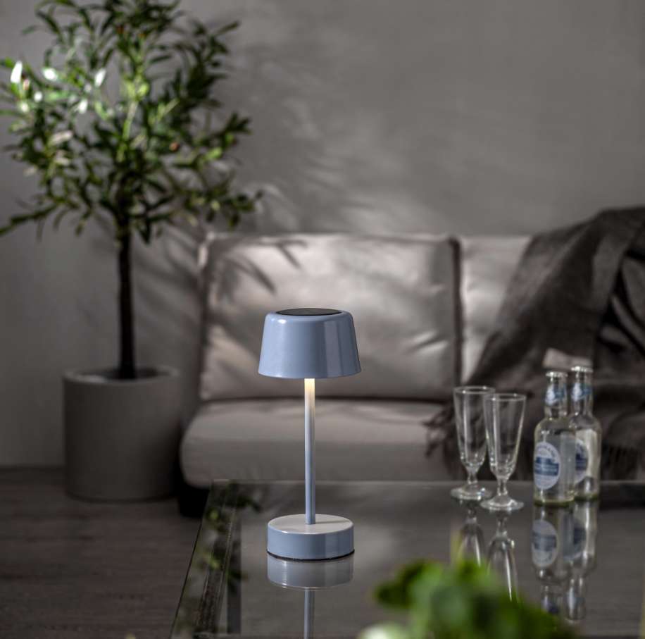 En tänd ljusblå sladdlös LED-bordslampa som skapar stämningsbelysning på ett glasbord i ett dämpat belyst, modernt vardagsrum.