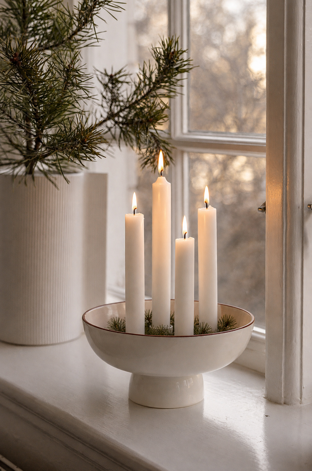 Vit adventsljusstake i keramik (Granhammar Jubileum, Storefactory) med tända ljus på en fönsterbräda bredvid en vit vas.