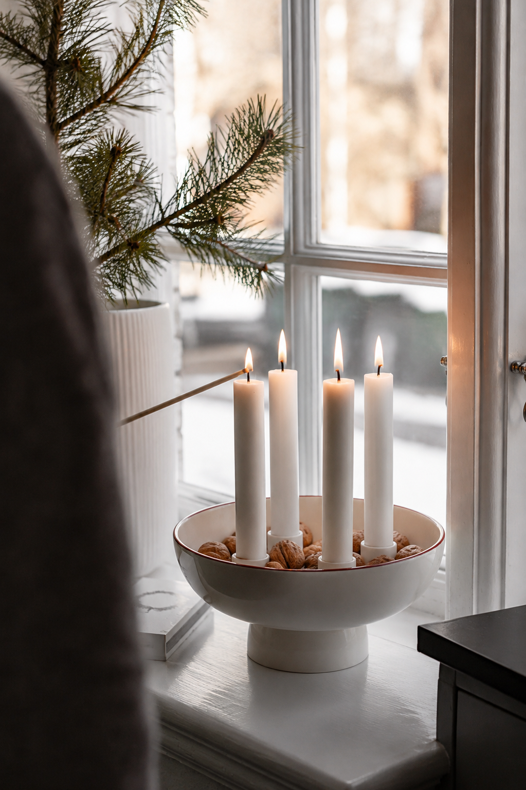 En person tänder första ljuset i adventsljusstaken Granhammar Jubileum från Storefactory. Den vita keramikskålen med röd kant är fylld med valnötter framför ett vintrigt fönster.