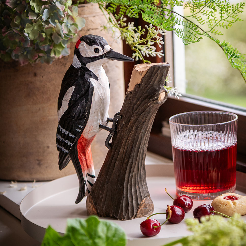 DecoBird Pic Épeiche sur un tronc d’arbre, entouré de plantes vertes, de cerises et d’un verre de boisson rouge.