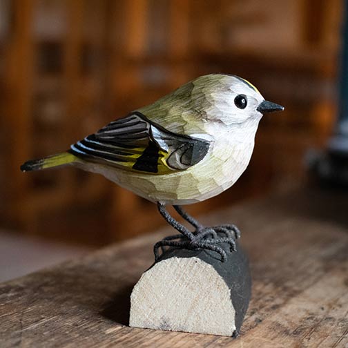 DecoBird Kungsfågel på träfot, placerad på ett träbord i en varm interiörmiljö.