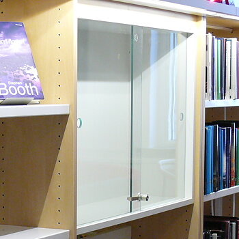 Vitrineskap/monter med låsbare skyvedører i glass og én glasshylle, integrert i hylleseksjon og tilpasset BK Librarys hyllesystem.