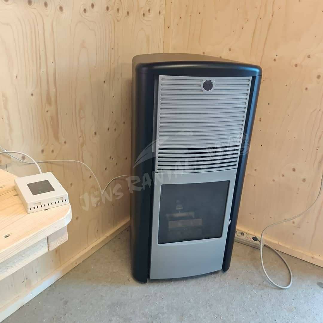 Skorstensfri pelletskamin Lilla Frö svart med grå front. Pelletskamin utan skorsten. I en verkstad. Monterad i ett hörn. Plywood på väggarna.