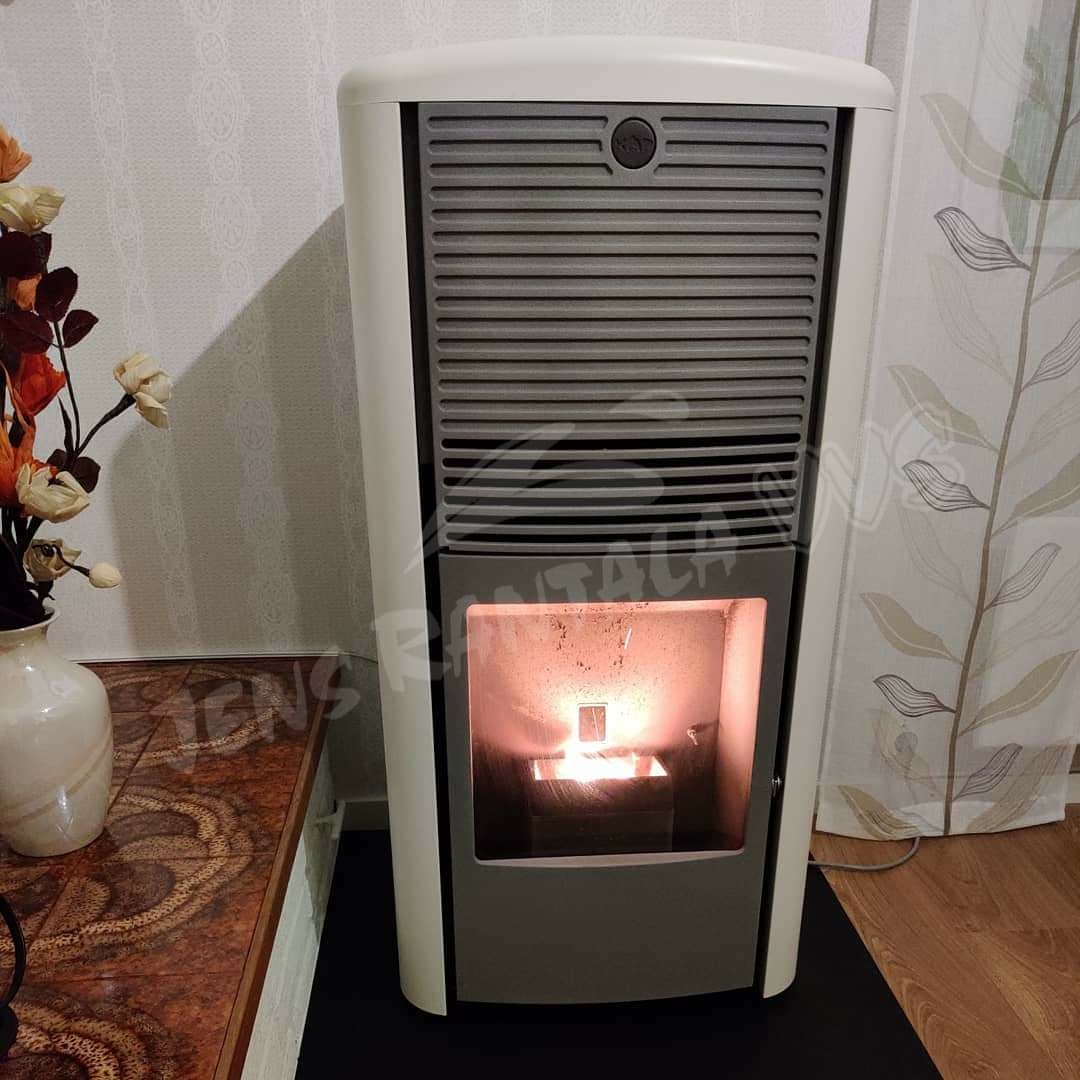 Skorstensfri pelletskamin Lilla Frö, vit med grå front. Pelletskamin utan skorsten. Här med vit gardin och hylla intill.
