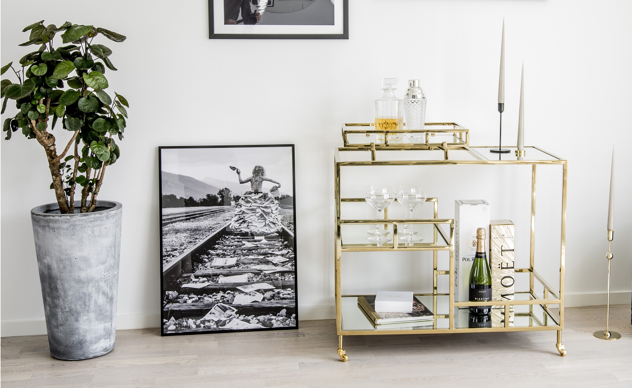 Drinkvagn i guld med champagneflaskor och whiskeyglas stående mot en vit vägg med svartvita posters och gröna växter