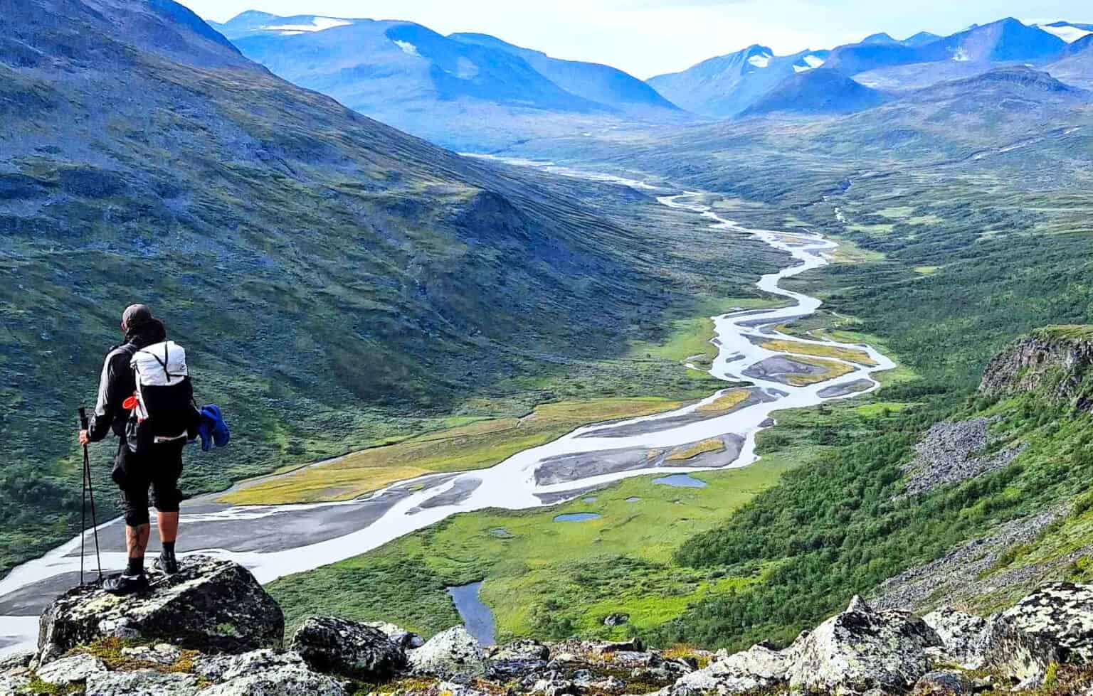 A hiker with a backpack and trekking poles stands on a rock looking out over a mountain valley with a braided river