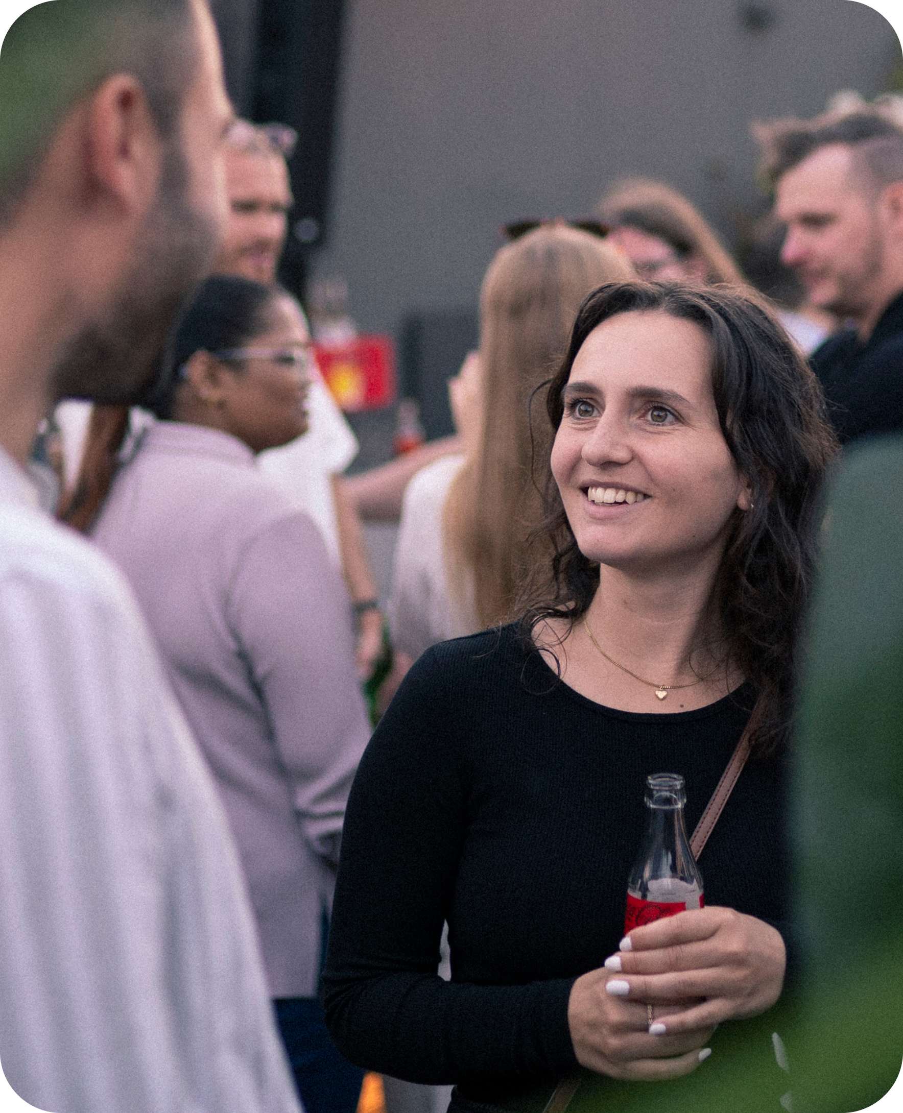 Kvinna som håller en flaska och ler i samtal med en man, med flera personer i bakgrunden på ett event.