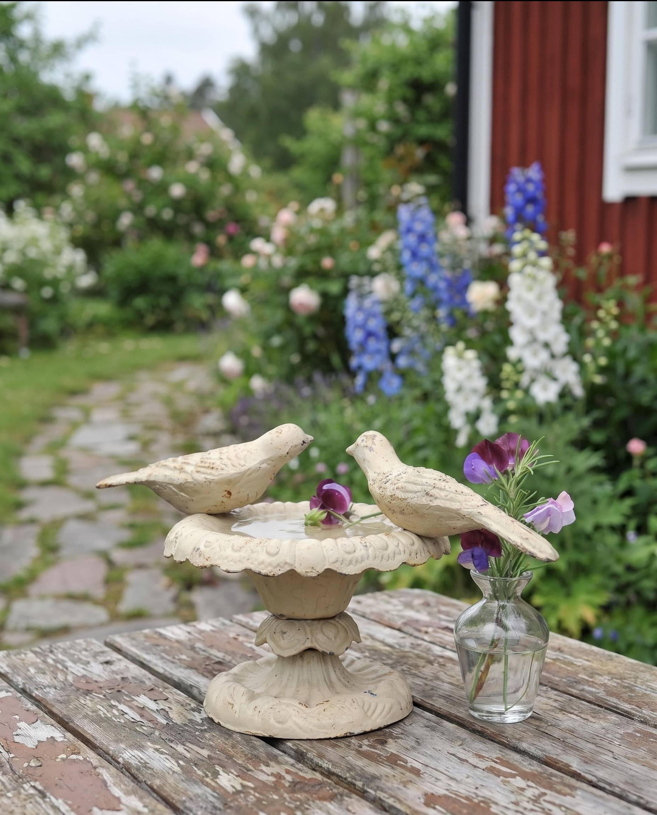 A vintage off-white cast iron bird bath with two perched birds and floating sweet peas, next to a clear glass vase with rosemary and flowers, sitting on a weathered wooden garden table in an authentic Swedish country garden.