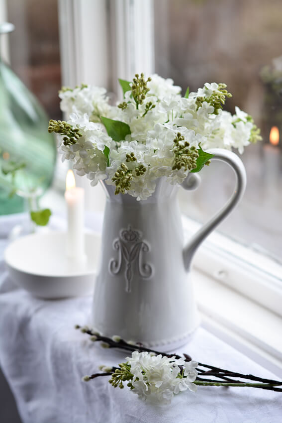 Artificial white lilac in white ceramic pot with M monogram in window, with willow twigs, linen cloth and lit candle
